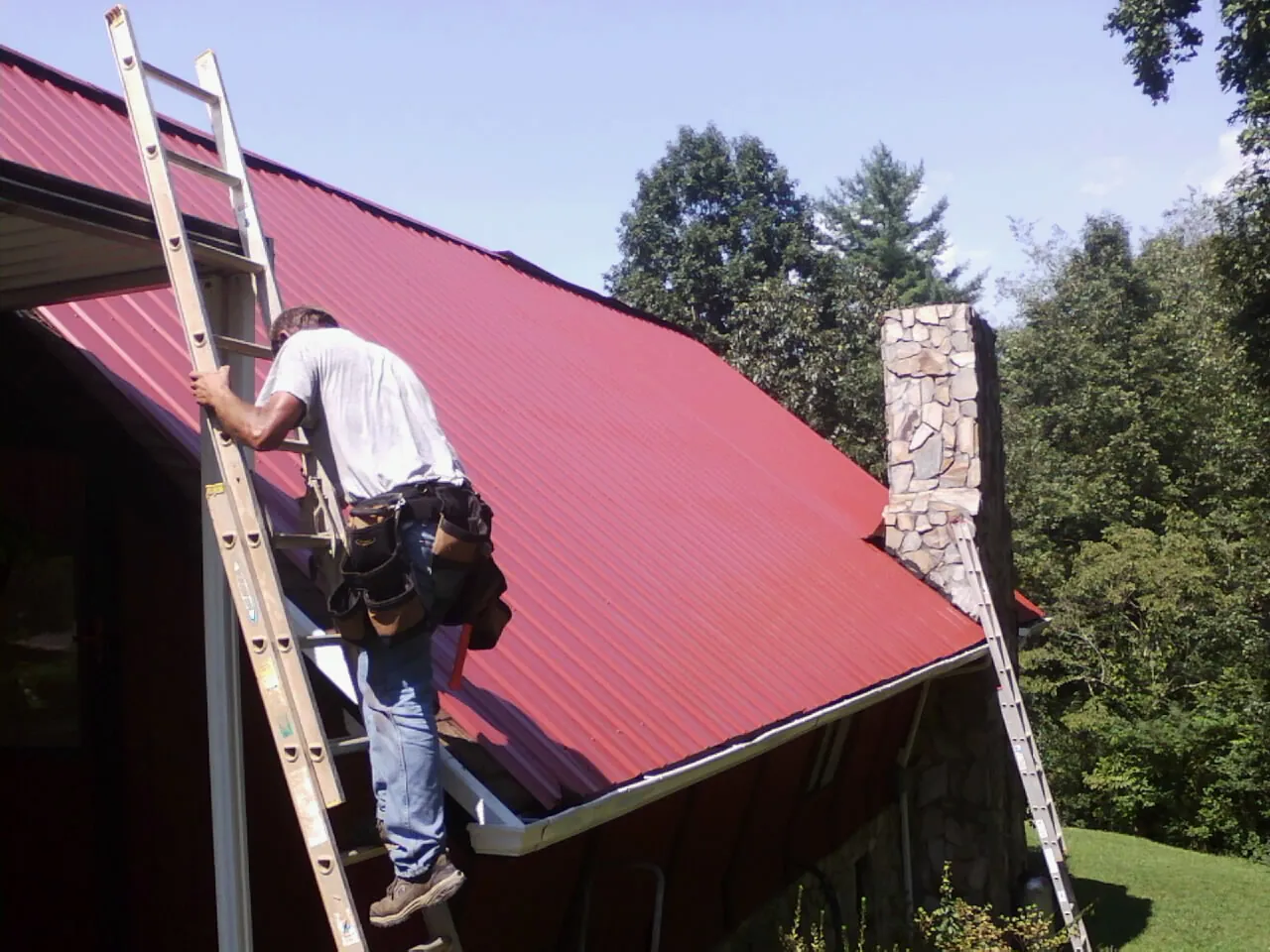 Roofer on a ladder beside a red metal roof and stone chimney — Soffit & Fascia Repair by Roofing Brothers in Tuxedo Park